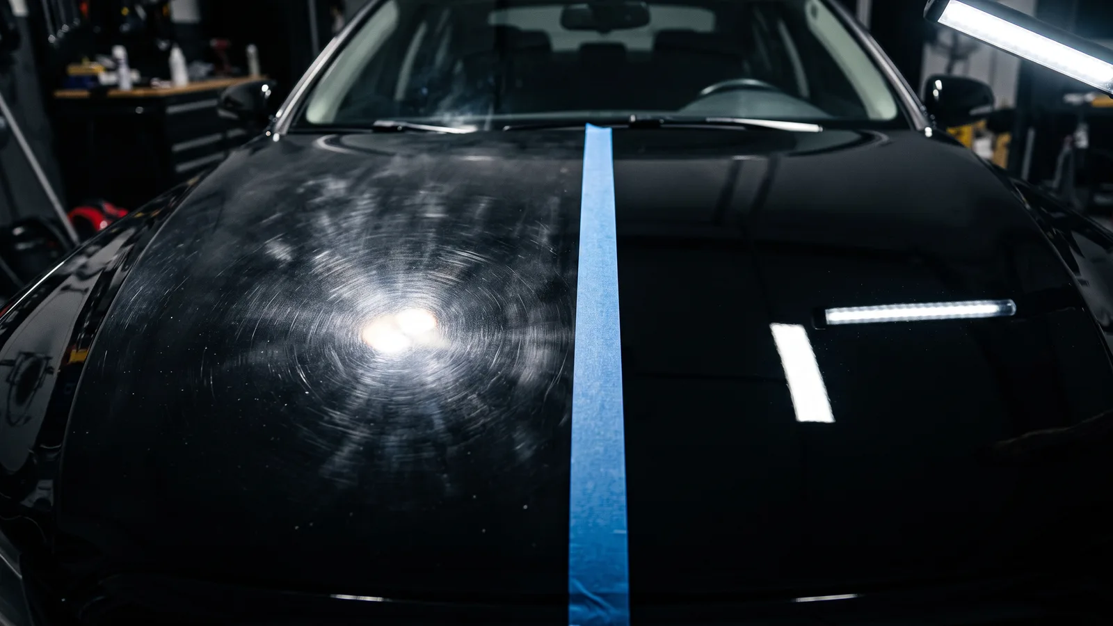 Black car hood split by masking tape under an LED inspection light, swirl marks on one side and polished mirror-like paint on the other, showing paint correction before selling