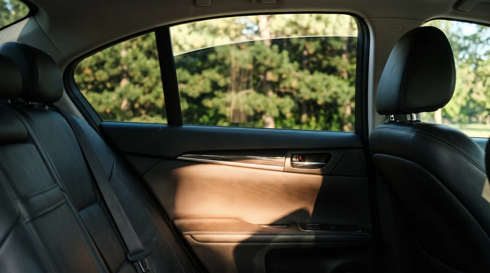 Rear passenger window with a light neutral window tint and a patch of sunlight on the door panel, trees outside still clearly visible, illustrating how a light legal tint can still reject infrared heat