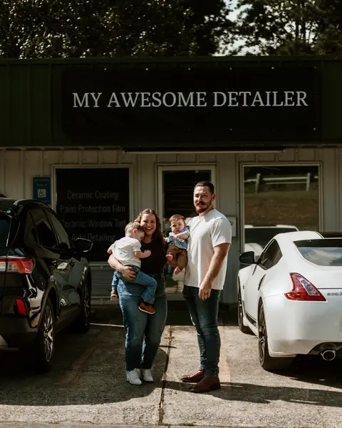 Christian Turner with his wife and their two young sons outside the My Awesome Detailer shop in Opelika, under the shop sign, beside his white 370Z